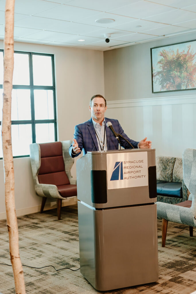 A person at a podium speaks at a Syracuse Regional Airport Authority event, surrounded by chairs and abstract artwork on the wall.