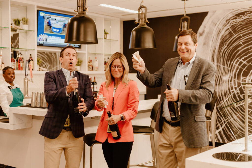 Three people celebrating in a modern indoor setting with champagne. A television is mounted on the wall, and a person observes nearby.