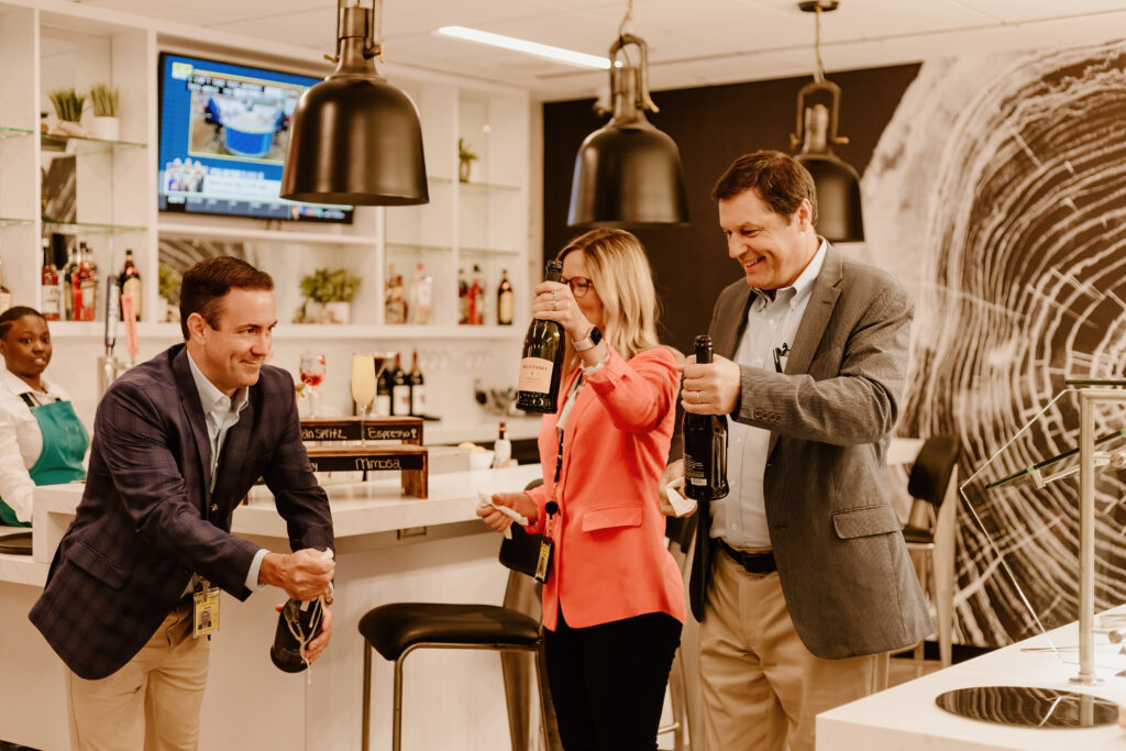Three people in a modern bar setting, holding bottles and glasses, smiling. A person in an apron stands nearby. Wall art visible.
