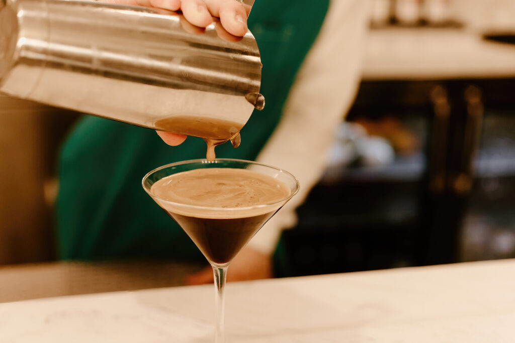 A person is pouring a chocolate-colored drink from a metal shaker into a martini glass on a white counter.