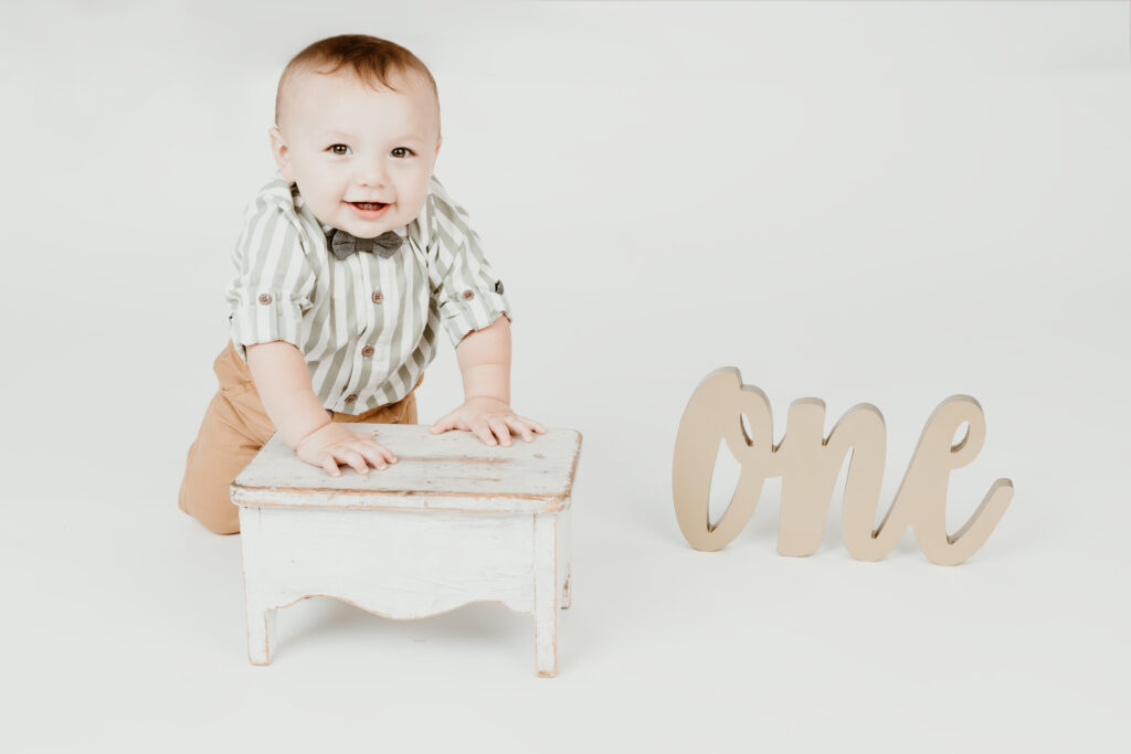 A smiling baby stands leaning on a white stool, wearing a striped shirt, bowtie, and tan pants. A wooden