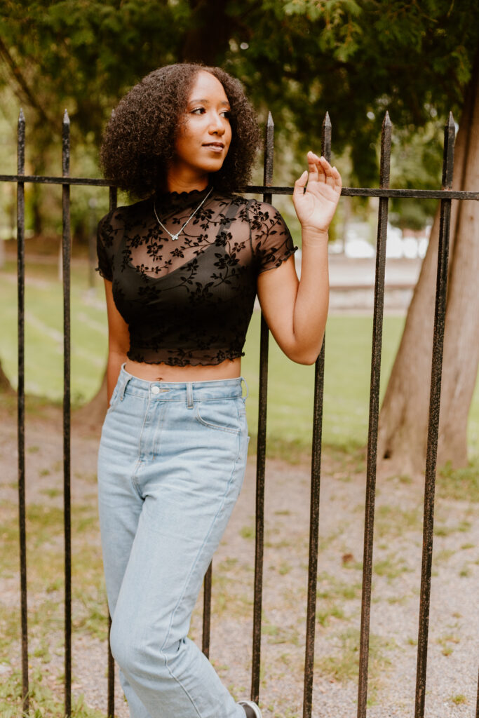 A person stands casually touching an iron fence, wearing a black lace top, light blue jeans, and sneakers, with trees in the background.