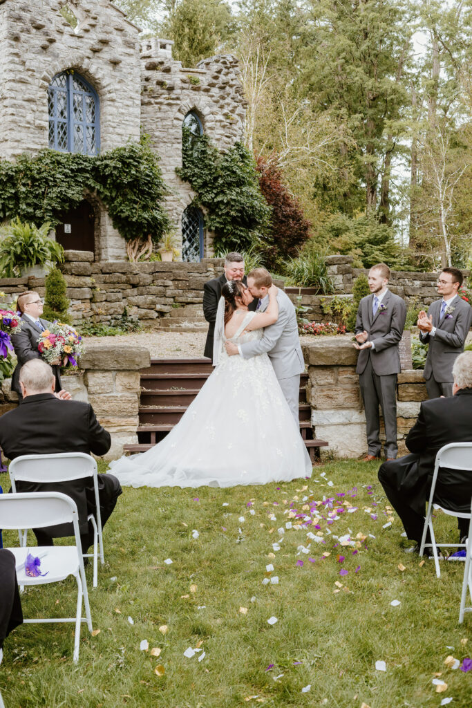 Two people are embracing at an outdoor wedding ceremony by a stone building, with guests and scattered flower petals around, exuding joy and celebration.