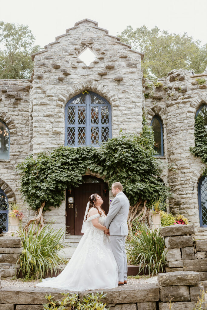 Two people, likely newlyweds, embrace in front of an old stone building with ivy, large windows, and a wooden door, conveying a romantic setting.