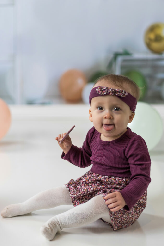 A baby with a purple headband and sweater sits on the floor holding a biscuit, with defocused balloons and light background.