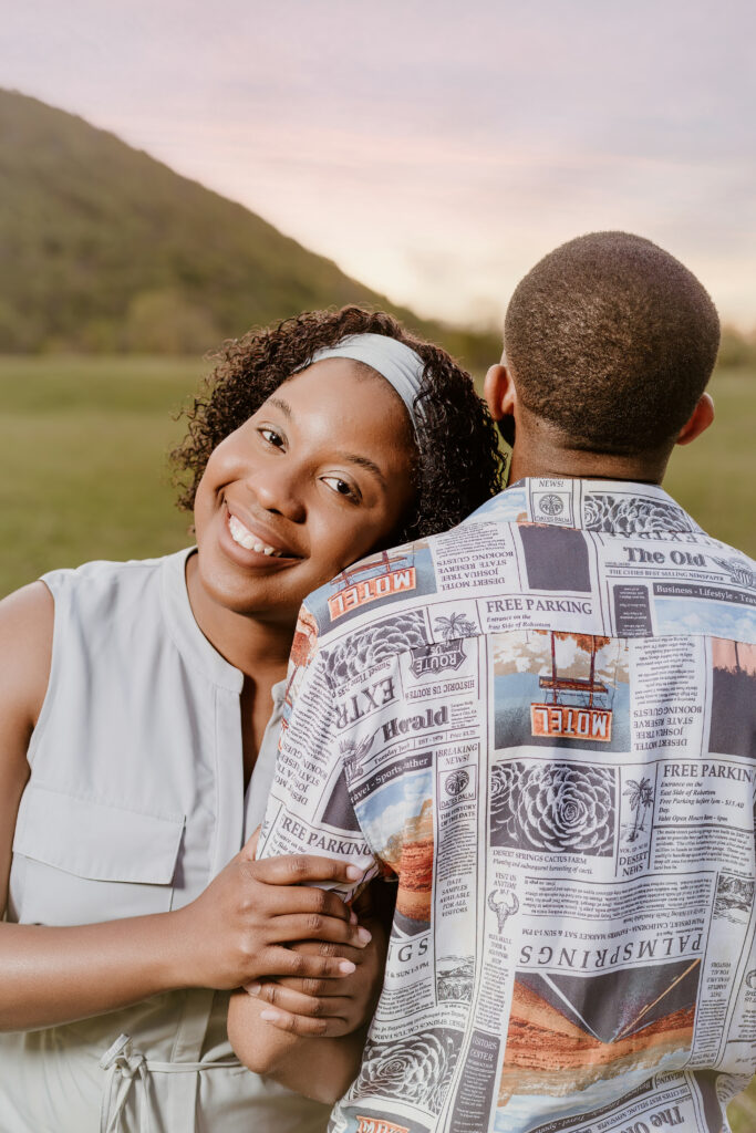 A person is engaging with a smiling person hiding behind a newspaper. They are outdoors with greenery and a soft pinkish sky in the background.
