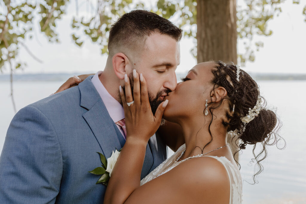 Two people share a kiss, dressed elegantly, likely a wedding scene. A lake and trees are visible in the background. It's a tender, romantic moment.