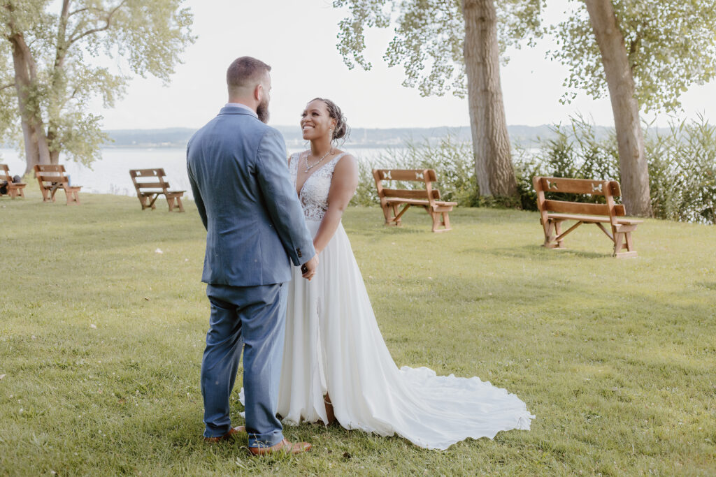 Two people in wedding attire are standing on a grassy lakeside. The person in a gown is smiling at the person in a suit. Trees and benches visible.