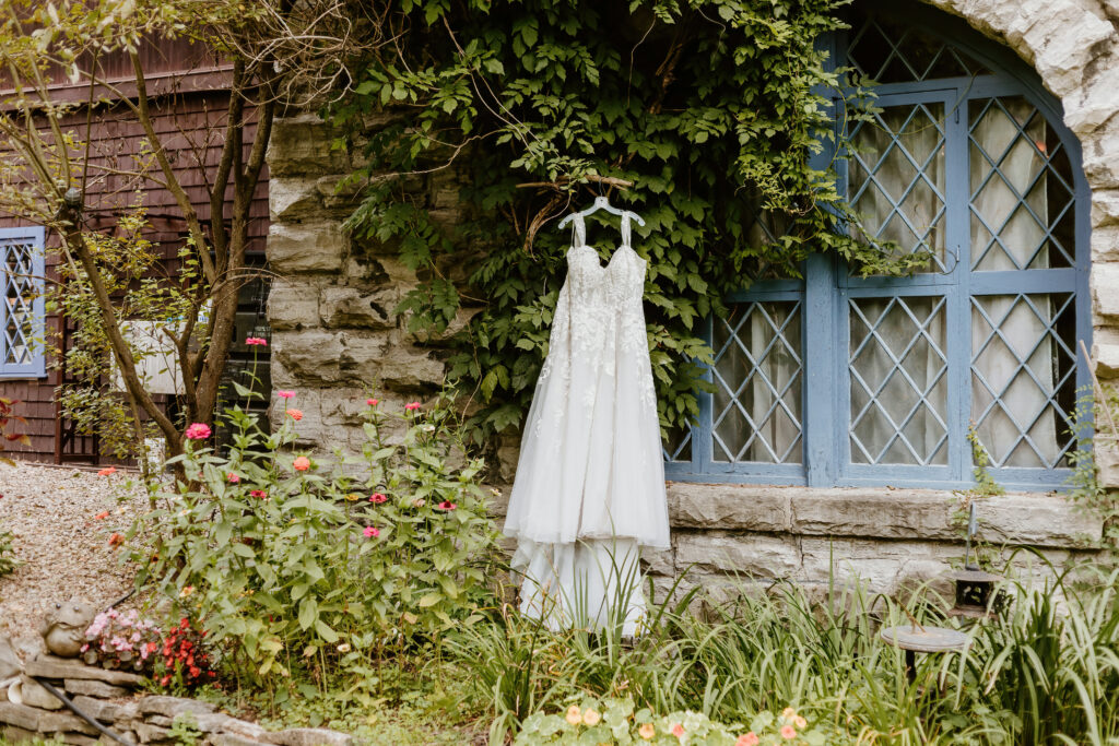 A bridal gown hangs beside a rustic building with blue windows, surrounded by greenery and flowers, creating a picturesque, serene, and romantic setting.