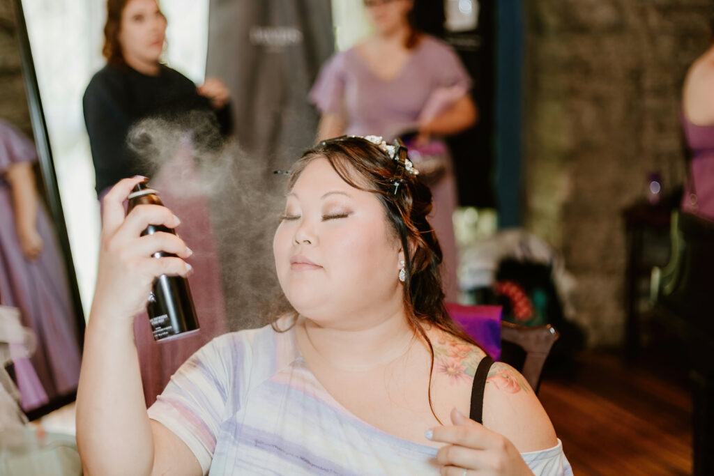 A person with closed eyes is spraying hair product, creating a mist around their face. They have a floral tattoo and elegant earrings. Others are in the background.