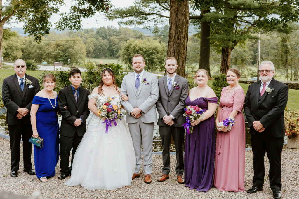 A group of eight people, dressed formally, stand outdoors with a background of trees and a grassy area, likely at a wedding event.