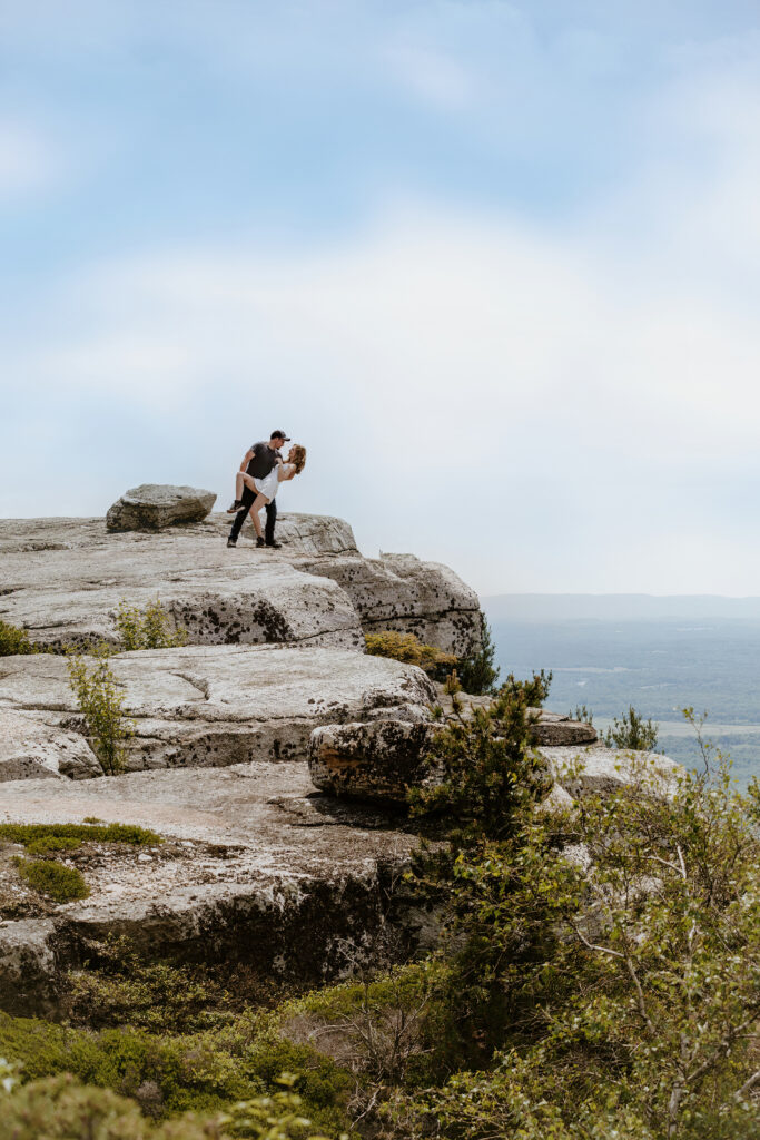 Two people embrace at the edge of a rocky cliff overlooking a vast landscape. It's a serene scene with clear skies and greenery below.