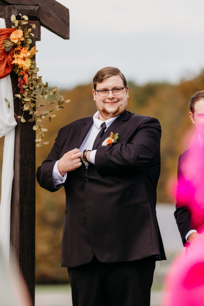 A man in a dark suit and boutonnière adjusts his watch at an outdoor wedding ceremony beside a flower-adorned wooden arch.