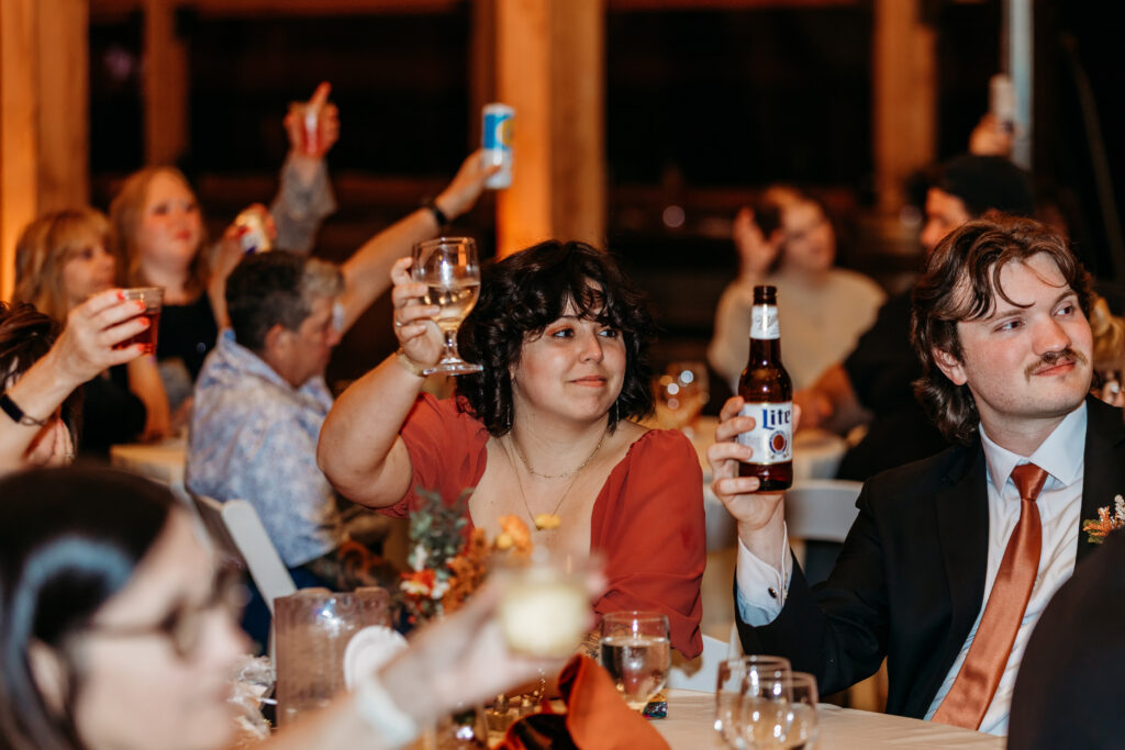 Guests at a banquet cheer with drinks, raising glasses and bottles around a long table.