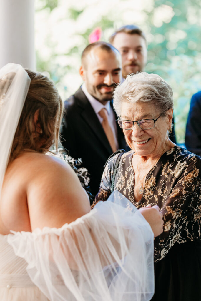An elderly woman with glasses in a patterned blouse smiles at the bride in a white wedding dress and veil during a wedding ceremony.