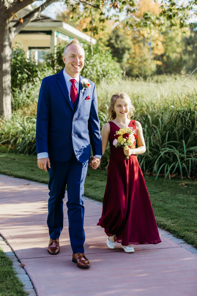 A man in a blue suit and a girl in a burgundy dress walk hand in hand along a pink path outdoors, with greenery in the background.