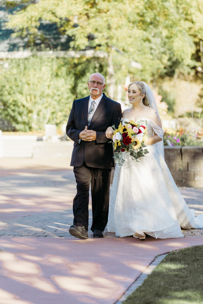 Bride in a white wedding dress walks arm-in-arm with an older man in a dark suit along a sunlit garden path.