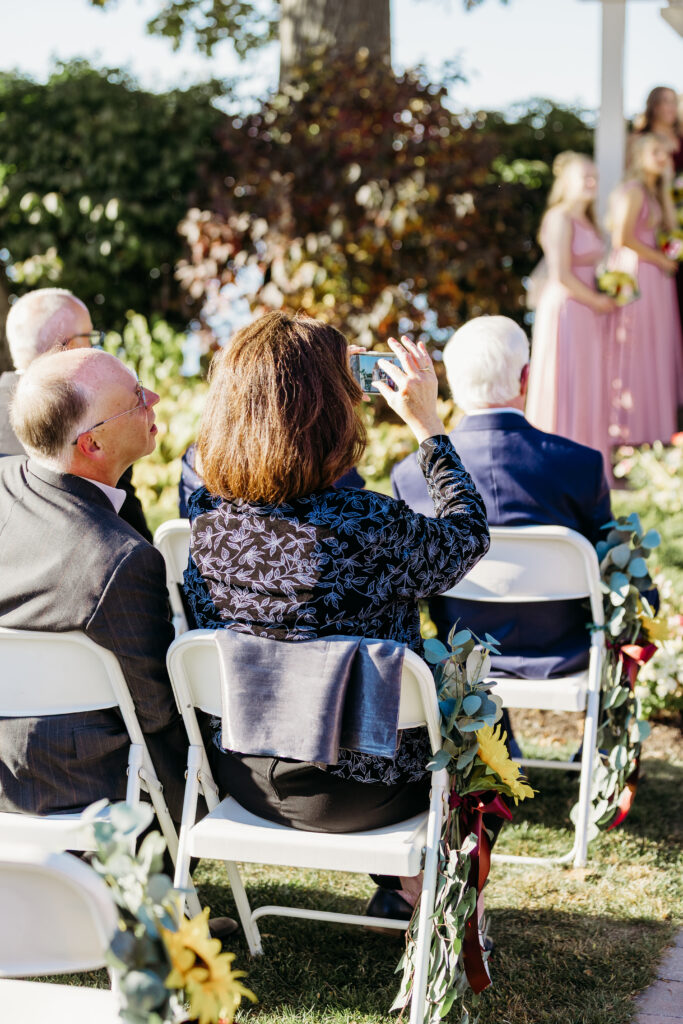 Outdoor wedding ceremony with guests seated on white chairs, floral aisle decorations, and two bridesmaids in pink dresses standing at the front.