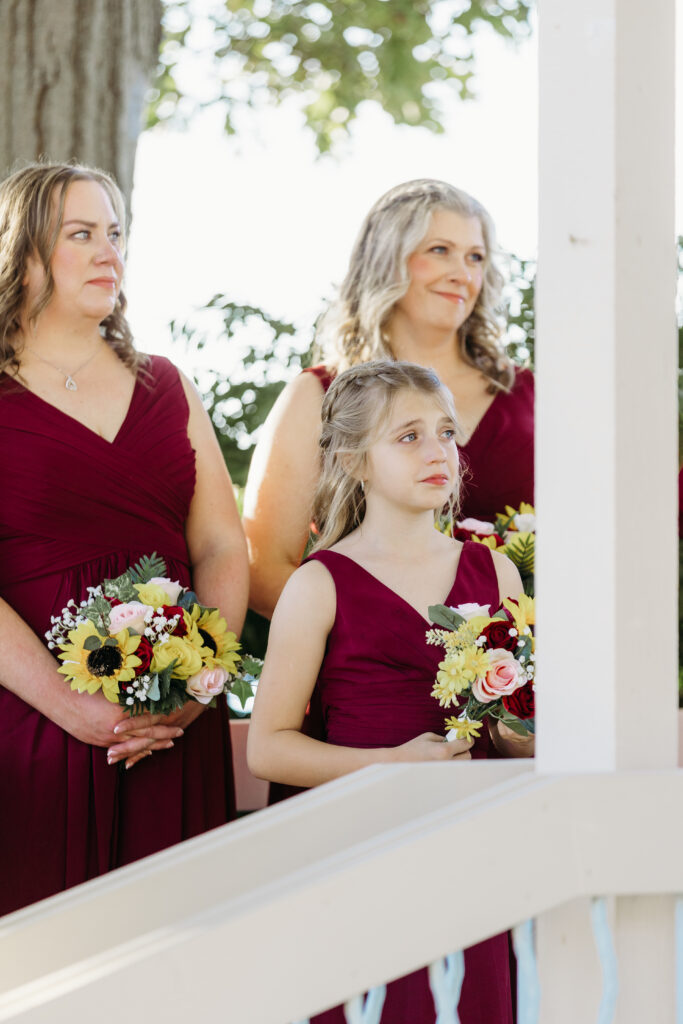 Three people in burgundy dresses hold bouquets on a white porch.