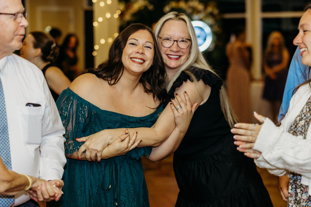 Two smiling women hug at a festive party, one wearing a teal off-shoulder dress and the other in black, with guests in the background.