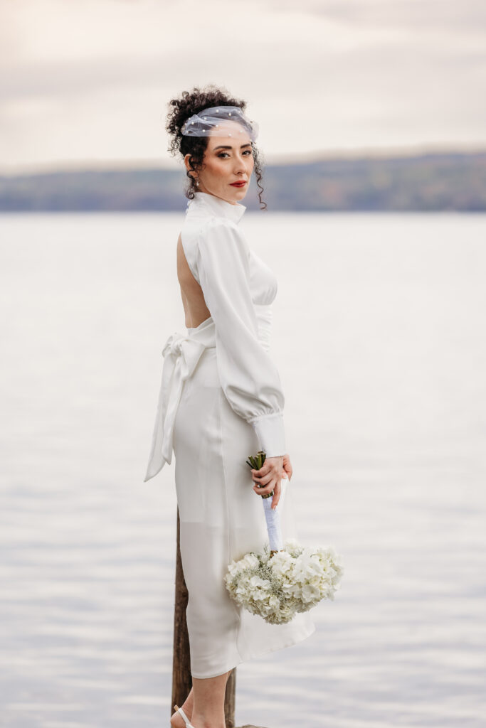 A woman in a white dress with an open back and side slit stands by a lake, holding a small bouquet of white flowers and looking over her shoulder.