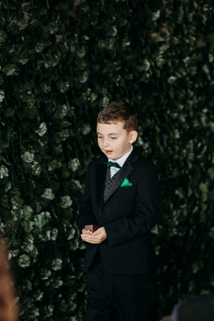 A young boy in a black suit with a bow tie and a green pocket square stands with his hands clasped in front of a leafy hedge.