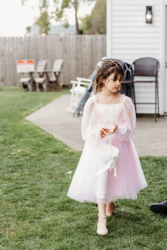 A young girl in a pink and white tutu dress walks across the lawn, holding a small bouquet in a backyard.