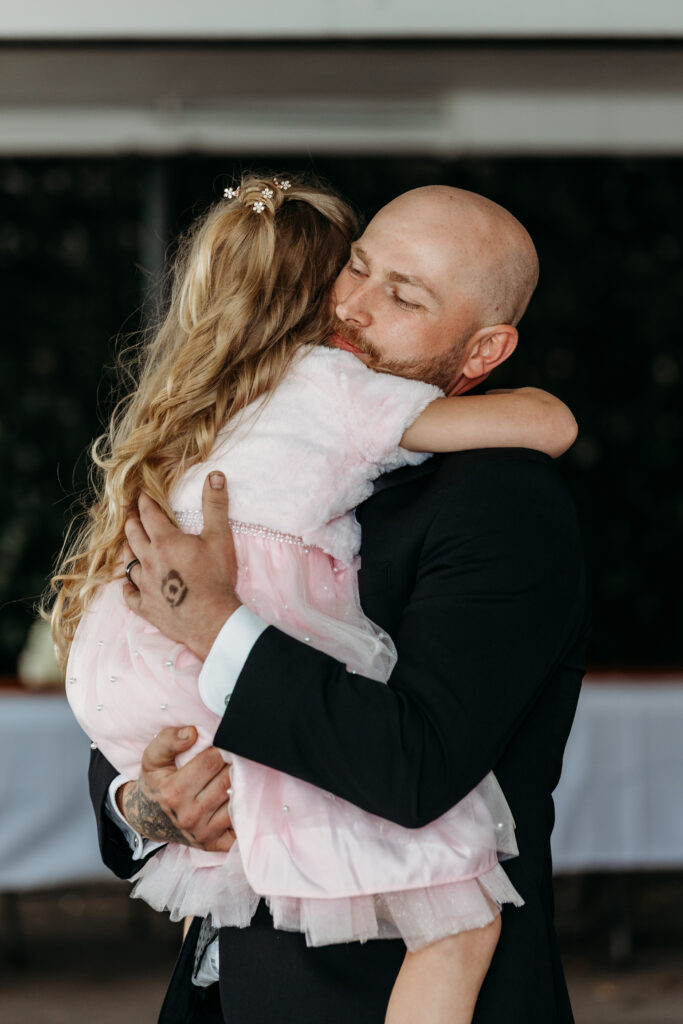 A man in a dark suit embraces a young girl in a pink dress.
