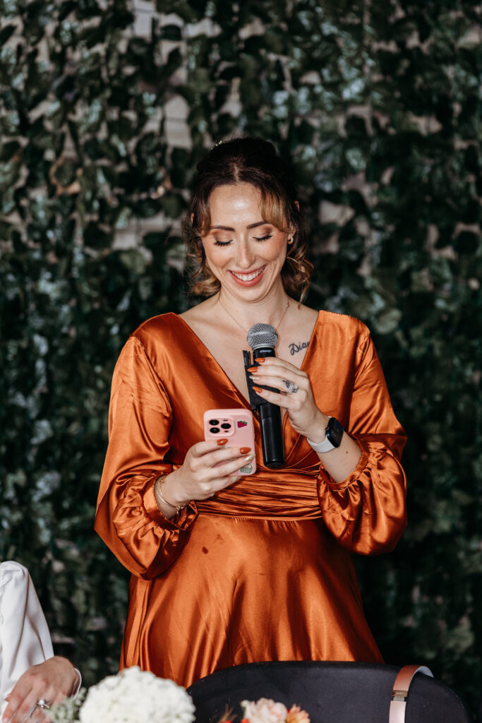 A smiling woman in an orange satin dress holds a microphone and pink smartphone on a stage with a leafy backdrop.
