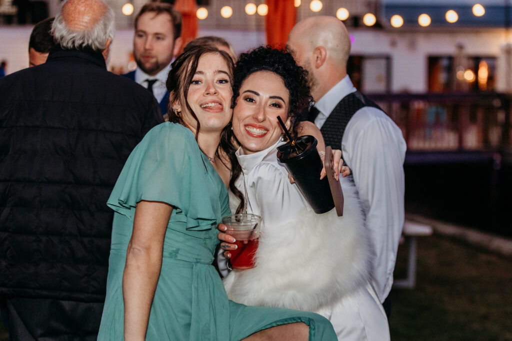 Two women posing at an outdoor party, one in a green off-the-shoulder dress and the other in a white fur stole, both holding drinks.