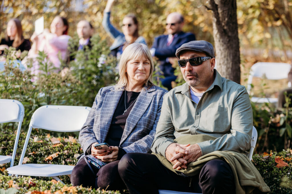 An older couple sits together on folding chairs in a park during autumn, with trees and other people in the background.