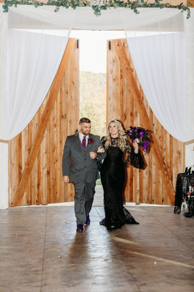 A couple in formal attire walks through an open wooden doorway with white curtains, toasting with glasses at an outdoor wedding.