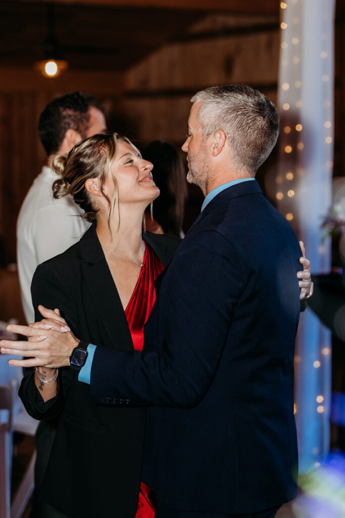 A couple dances closely at a formal event, smiling at each other with warm string lights in the background.