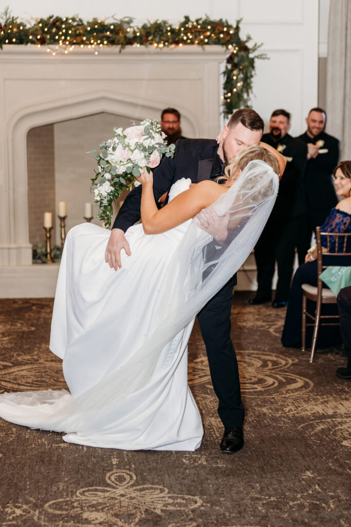 Groom lifts his bride in a white wedding dress during a dip at an indoor ceremony, with guests watching and festive garlands in the background.