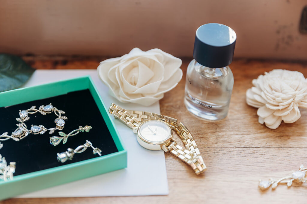 A gold wristwatch, a turquoise jewelry box with silver jewelry, a small perfume bottle, and white flowers on a wooden surface.