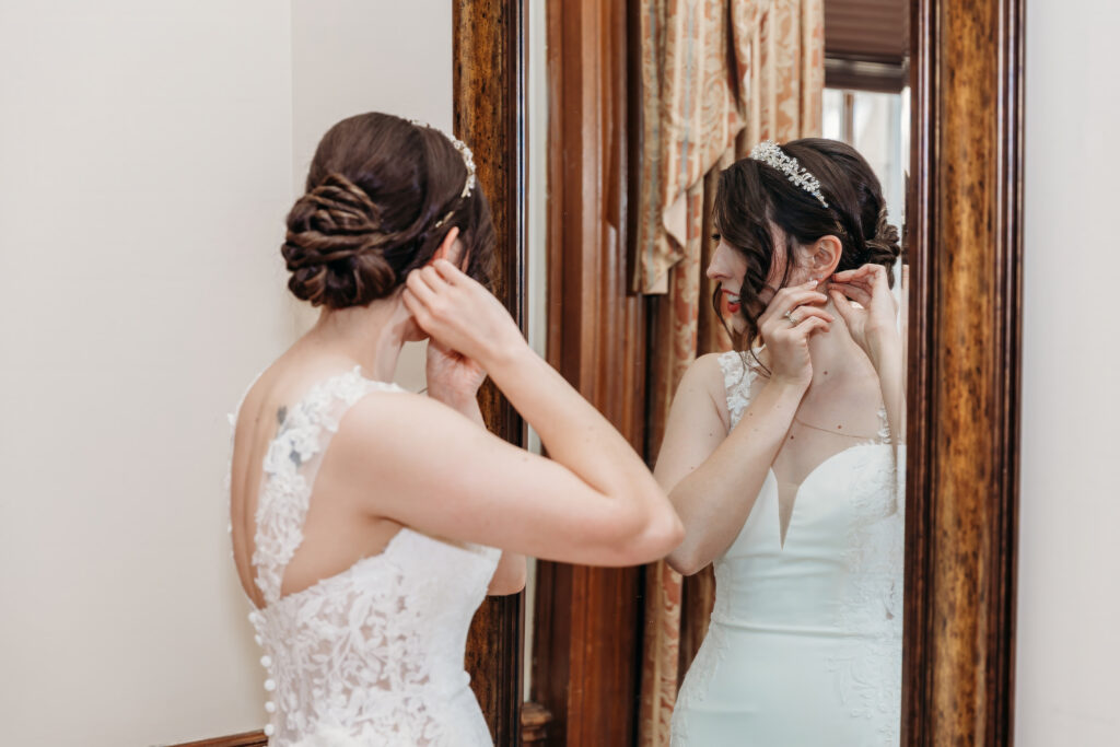 Bride in a white lace wedding dress adjusts her hair while looking in a wooden-framed mirror.