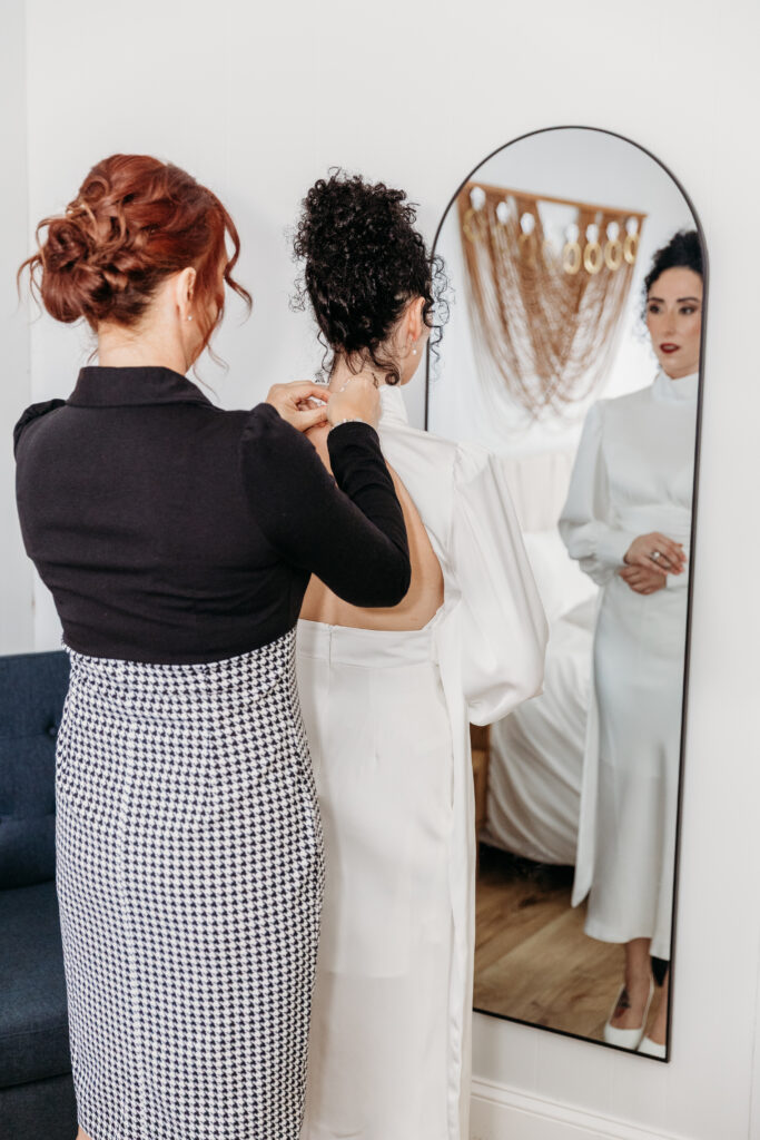 Three women in a bridal fitting room, with a bride in a white wedding gown being helped to adjust the dress, visible in a full-length mirror.