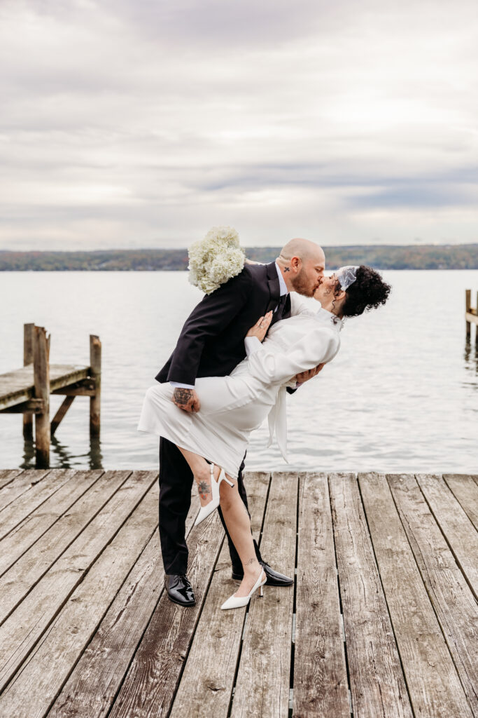 Bride and groom share a kiss as he dips her on a wooden dock by a lake.