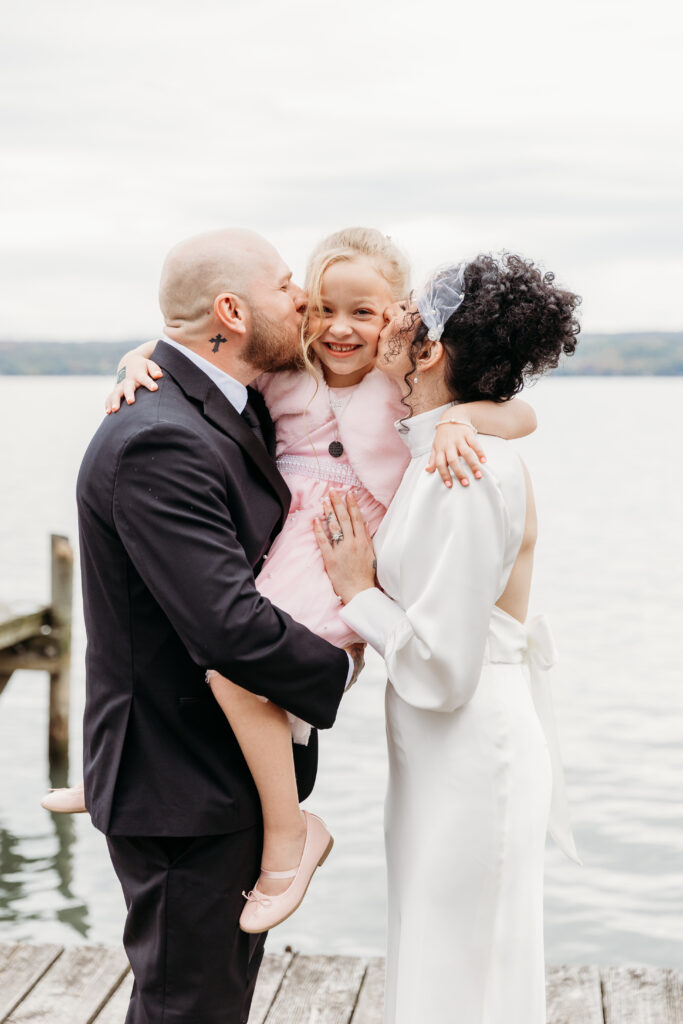 A bride and groom hug their smiling daughter on a wooden dock by the water.