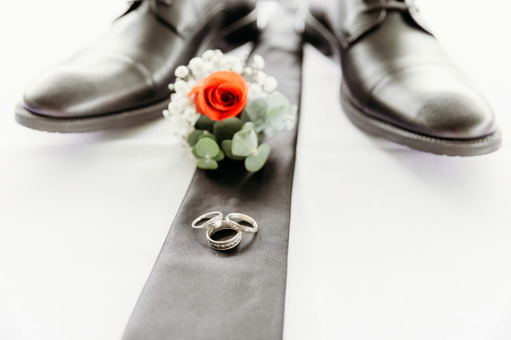 A pair of polished black dress shoes, a boutonniere with a red rose, and two wedding rings placed on a dark tie on a white background.