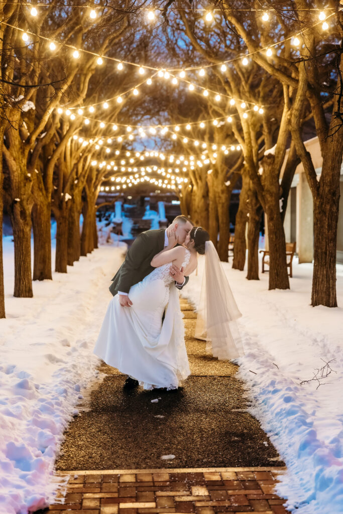 A couple walks along a snow-covered brick path lined with trees and twinkling string lights overhead.