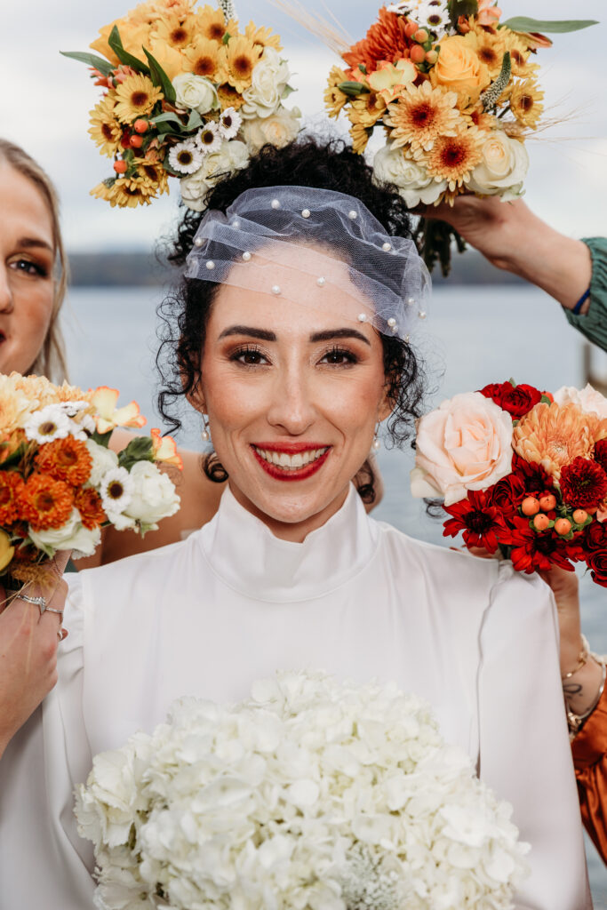 Bride in a white dress with veil and flower crown, smiling and holding a white bouquet while surrounded by bridesmaids with colorful bouquets.