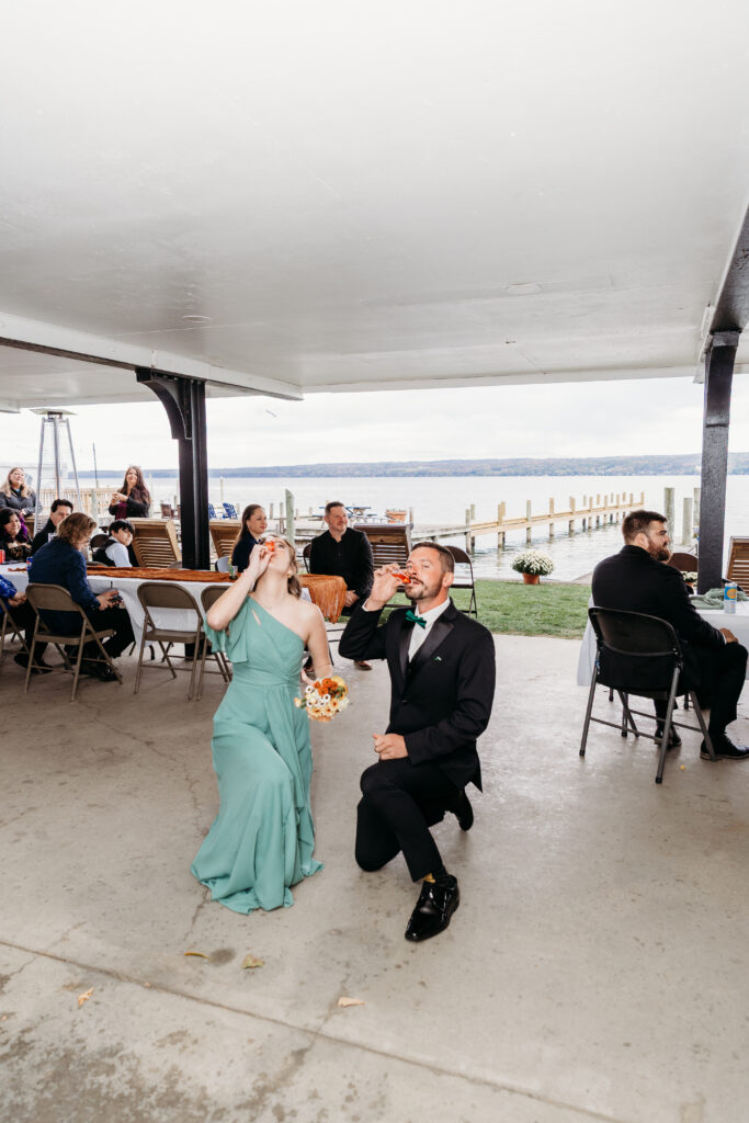 A man on one knee proposes to a woman in a teal gown at an outdoor waterfront wedding reception under a white canopy, with guests seated at tables in the background.