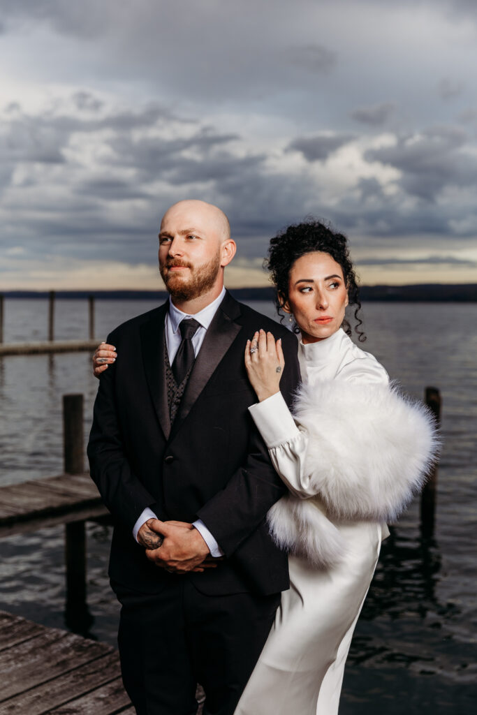 A couple stands on a wooden dock by a cloudy body of water, the man in a dark suit and the woman in a white fur-trimmed coat posing together.