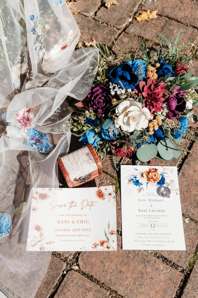 Colorful bouquet of blue, pink, and white flowers rests on a brick patio beside two wedding invitation cards and a small pink box, with sheer fabric nearby.