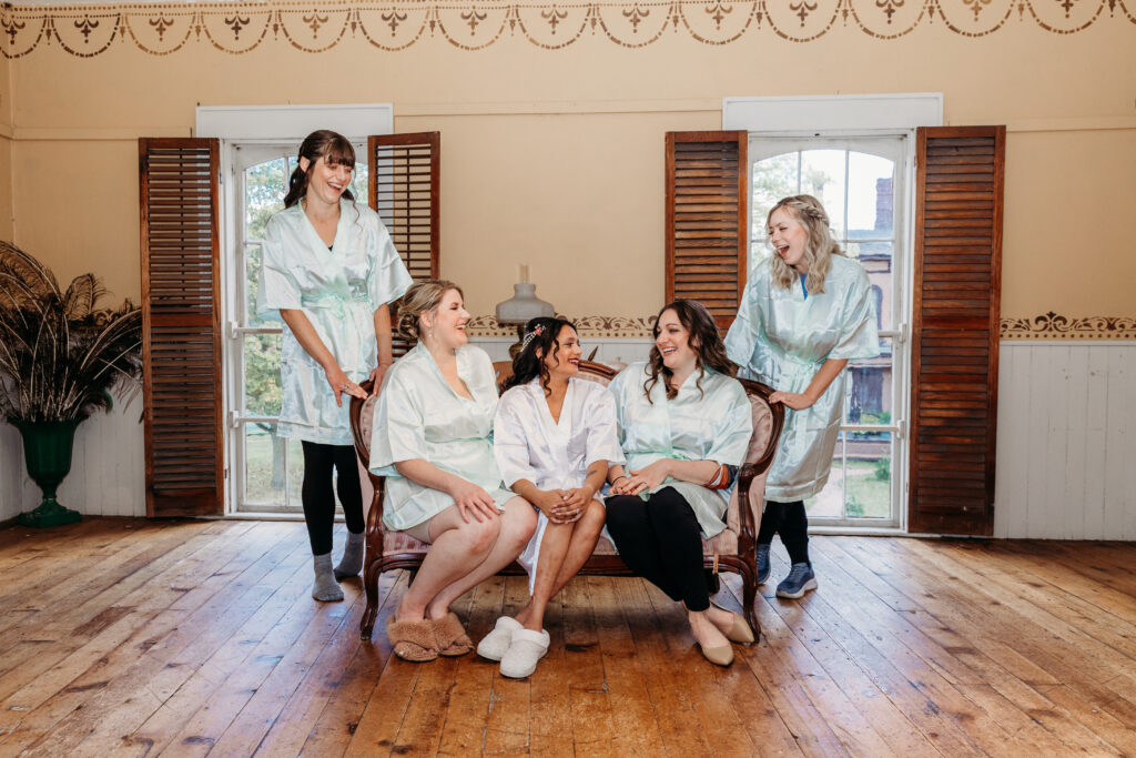Six women in light green robes pose together in a sunlit room with wooden floors and shuttered windows.
