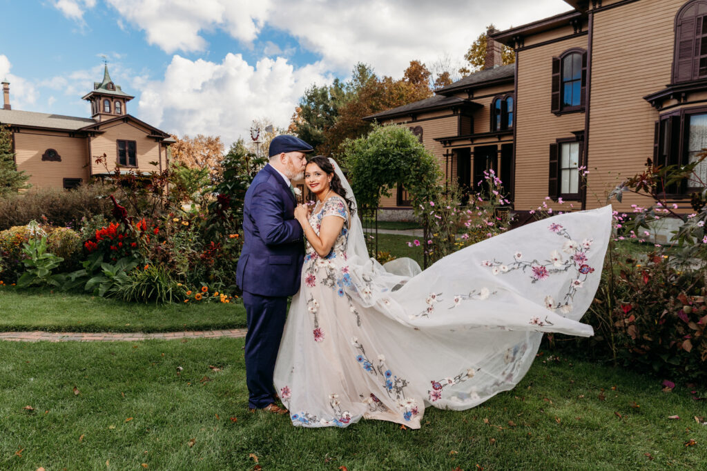 A bride in a flowing floral-embroidered wedding gown and a groom kiss on a grassy lawn in front of a brick mansion.