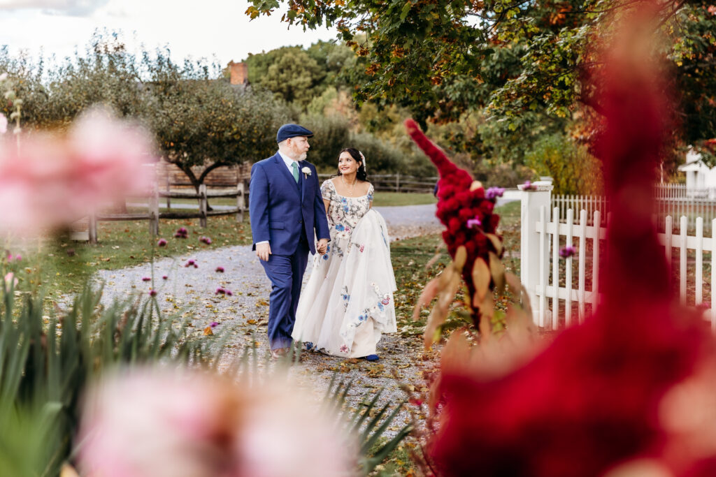 A bride and groom walk hand in hand along a garden path beside a white picket fence, surrounded by colorful blooms.