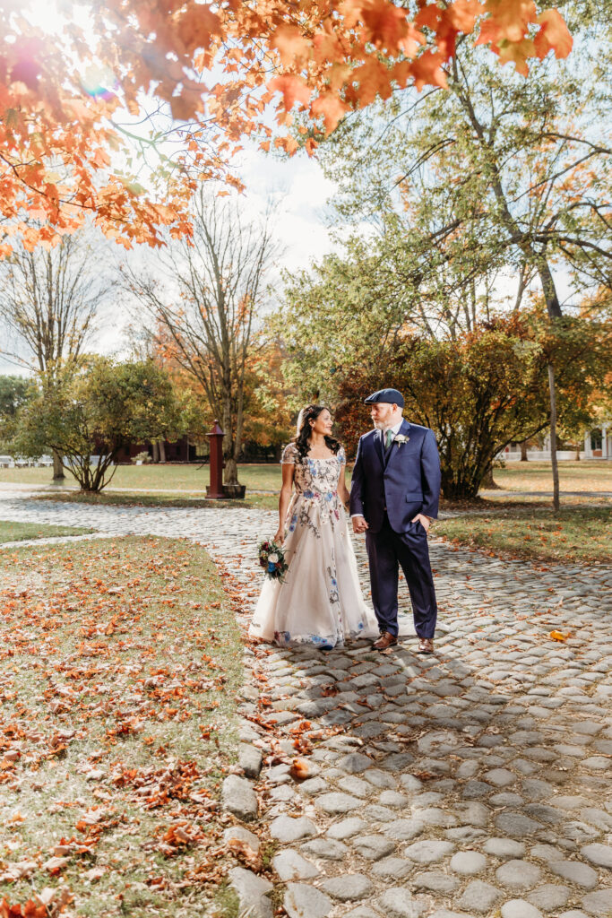 A couple in formal attire stands on a cobblestone path in a park during autumn, the woman in a light gown holding a bouquet as orange leaves fall from the trees.