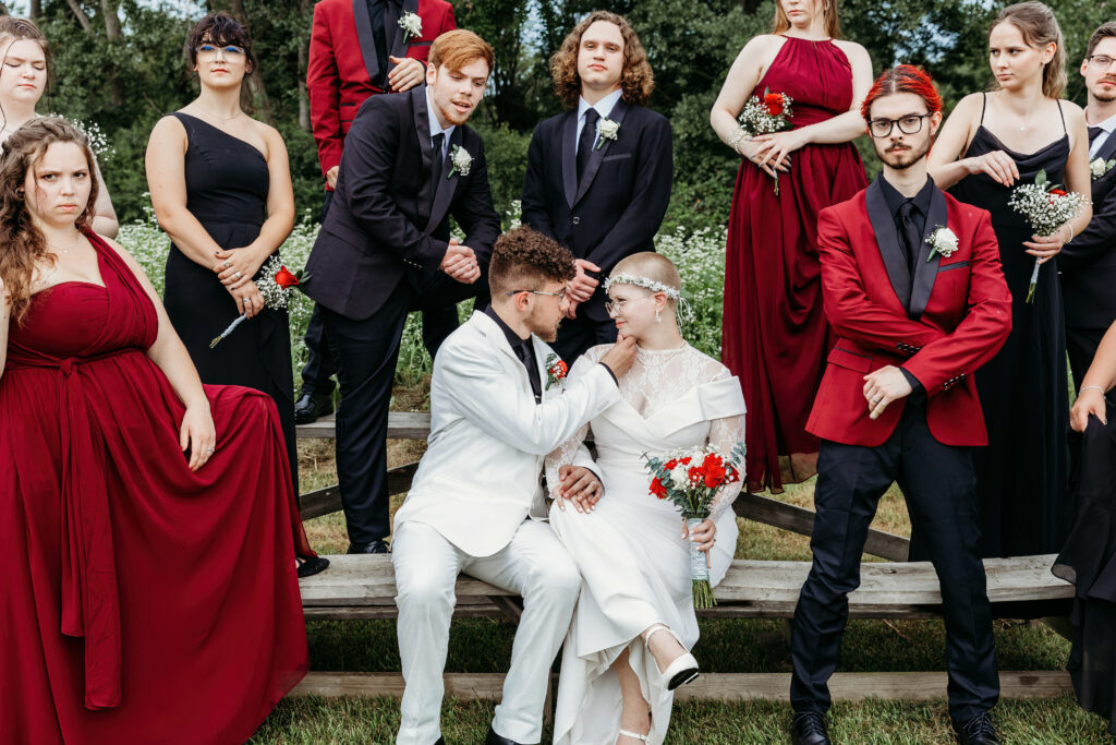 A group of people in formal attire poses outdoors. One person in white and another person sit in the middle with others standing around.